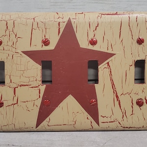 May include: Cream-coloured light switch cover with a distressed finish and a large red star in the centre. The cover has four switch openings and is secured with red screws. The design has a rustic, country aesthetic.