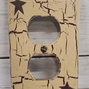 May include: Tan electrical outlet cover with a distressed finish and two dark brown star accents. The cover has two openings for outlets and a screw in the centre. The surface has a crackle effect.