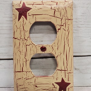 May include: A cream-coloured outlet cover with a distressed finish and red stars. The cover has two outlets and a screw in the centre.