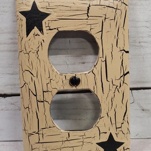 May include: Tan electrical outlet cover with a distressed, crackled finish and two black stars. The cover has two circular openings for the outlets and a single screw in the centre.