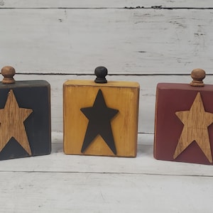 May include: Three wooden blocks in navy, yellow, and burgundy, each with a star cutout. The navy block has a natural wood star, the yellow block has a black star, and the burgundy block has a natural wood star. Each block has a small wooden knob on top.