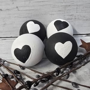 May include: Four round decorative objects, painted in black and white, each featuring a heart design. Two are black with white hearts, and two are white with black hearts. The objects are arranged on a rustic, light-colored wooden surface.