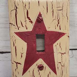 May include: A light switch cover with a distressed, cream-colored background and a large, red star painted in the center. The star has a light switch opening in the middle.