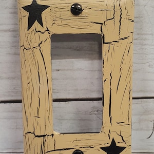 May include: A light beige light switch cover with a distressed woodgrain pattern and black stars. The cover has two black screws.