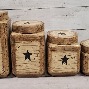May include: Set of four decorative wooden storage boxes with lids. The boxes are painted in a distressed beige finish with black star accents. The lids have a wood grain appearance.