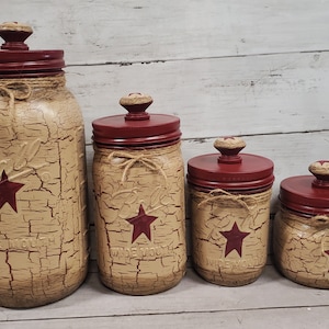 May include: Set of four rustic farmhouse style glass jars with red lids and star accents. The jars are painted in a distressed beige finish with a crackle effect. The lids have a red finish and a wooden knob.