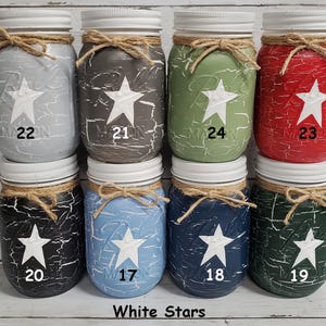 May include: Ten mason jars with white stars painted on them. The jars are painted in various colors, including light blue, dark blue, green, red, black, and gray. Each jar has a white star in the center and a number from 17 to 24 written below the star.