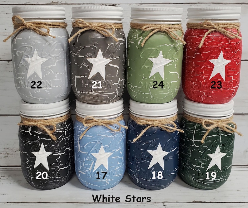 May include: Ten mason jars with white stars painted on them. The jars are painted in various colors, including light blue, dark blue, green, red, black, and gray. Each jar has a white star in the center and a number from 17 to 24 written below the star.