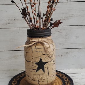 May include: A distressed cream-colored mason jar with a black star and the word "MASON" painted on it. The jar is filled with dried brown and black twigs and sits on a black and brown woven round mat.
