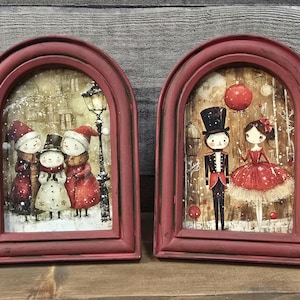 May include: Two arched, distressed red wooden frames containing holiday-themed illustrations. The left frame shows three snowmen in a snowy street scene with a lamp post. The right frame depicts a nutcracker and a ballerina in a red dress.