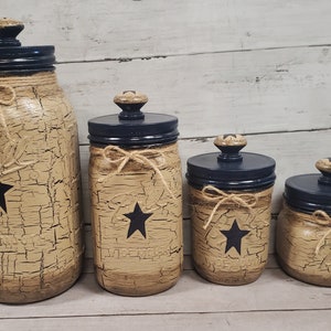 May include: Set of four rustic farmhouse style glass jars with black lids and twine accents. Each jar features a black star design and a distressed beige finish.
