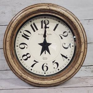 May include: A round, cream-colored clock with a distressed, aged finish. The clock face features black Roman numerals and a black star in the center. The clock is framed by a thick, brown wooden ring. The clock face reads "Sterling Noble Clock Company".