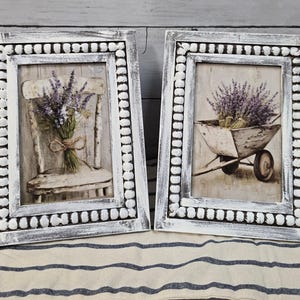 Puede incluir: Dos cuadros enmarcados. Uno presenta un ramo de lavanda sobre una silla blanca, el otro una carretilla llena de lavanda. Ambos están en marcos blancos con un borde de cuentas, creando una estética rústica de granja.