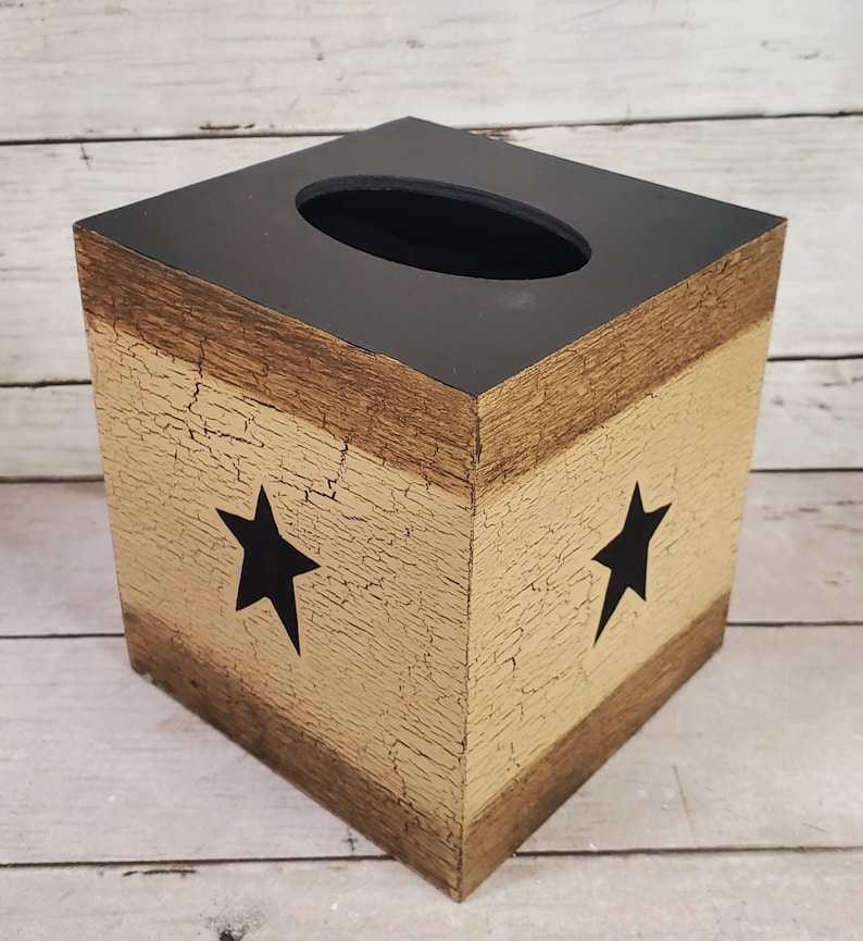 May include: A square tissue box cover made of wood with a distressed finish. The box is painted in shades of brown and beige with black stars on each side.