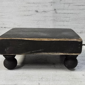 May include: A rectangular wooden riser with a distressed black finish and visible natural wood edges. The platform is supported by four small, rounded, black ball-shaped feet, providing a low-profile elevation for the flat, solid surface.