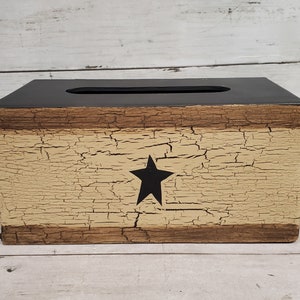 May include: A rustic wooden tissue box cover with a distressed beige and brown finish. The cover features a black star in the center.
