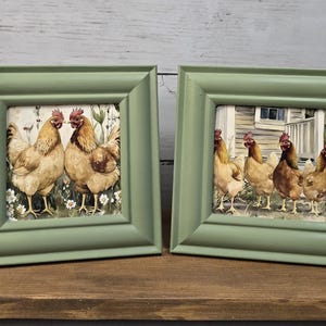 Può includere: Due cornici quadrate color verde salvia con illustrazioni di galline. La cornice a sinistra mostra due galline marroni tra fiori bianchi. La cornice a destra mostra quattro galline marroni davanti a una struttura in legno di colore chiaro.