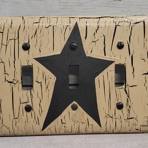 May include: A beige light switch cover with a black star design. The cover has a distressed, cracked paint finish.