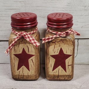 May include: Two rustic salt and pepper shakers with red lids and a red star design on a beige background. The shakers are tied with a red and white gingham ribbon.