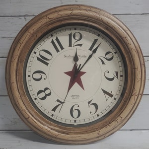 May include: A round, cream-colored wall clock with a distressed finish. The clock face has black Roman numerals and a red star in the center. The clock is made by Sterling Noble Clock Company.