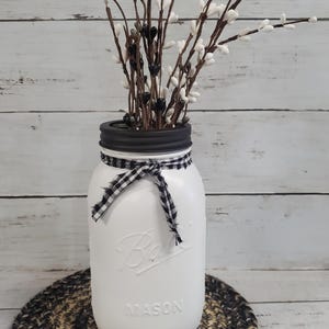 May include: White painted mason jar with a black and white checkered ribbon tied around the lid. The jar is filled with white and black faux pussy willow branches and is sitting on a black and tan woven round mat.