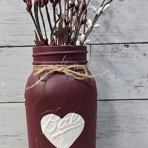 May include: A burgundy painted Mason jar vase with a white heart design and twine tied around the rim. The vase is filled with decorative branches with red berries, white berries, and rusty star accents. The word "MASON" is visible on the jar.
