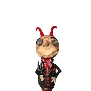 Può includere: Una piccola figura di diavolo dalla pelle rossa con corna, una giacca nera e pantaloni rossi. La figura tiene un forcone e ha un'espressione maliziosa sul viso. Il testo "DARTIAN DEVILISH" è scritto sotto la figura.