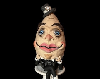 Peut inclure: Une figurine en céramique d'une personne avec une grosse tête en forme d'œuf, portant un smoking noir et un chapeau haut de forme avec un bijou. La figurine est assise sur un piédestal blanc et le mot "Henry" est écrit en lettres blanches sous le piédestal.