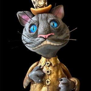 Könnte beinhalten: Skulptur einer Katze mit einem goldenen Jackett und einem karierten Zylinder mit einem Juwel. Die Katze hat leuchtend blaue Augen und eine rosa Nase, vor einem schwarzen Hintergrund. Die Katze grinst.