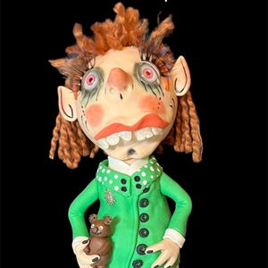 Può includere: Una bambola stravagante con capelli rossi, vestiti verdi e un orsacchiotto di peluche marrone. La bambola ha occhi grandi e un ampio sorriso. La bambola è etichettata "Penelope".