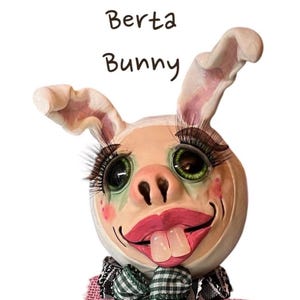 May include: A white bunny doll with large green eyes, pink lips, and a toothy grin. The doll is wearing a white bunny costume with long eyelashes and a pink bow tie. The doll is titled "Berta Bunny".