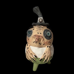 May include: A whimsical, hand-crafted clay figurine of a slug wearing a black hat with a silver button. The slug has large, blue eyes, a brown nose, and a green body with black spots.