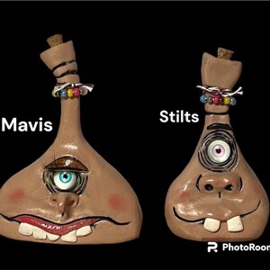 May include: Two ceramic bottle stoppers with whimsical faces. One has a blue eye and the other has a pink eye. Both have a single tooth sticking out of their mouths. The bottle stoppers are decorated with colorful beads and the words "Mavis" and "Stilts" are written on the image.