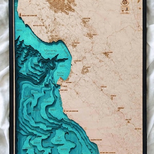 May include: A framed wooden map of the Monterey Bay area, featuring a detailed topographic representation. The map highlights the Monterey Canyon with varying depths in shades of teal and blue, set against a light brown landmass. The frame is black.