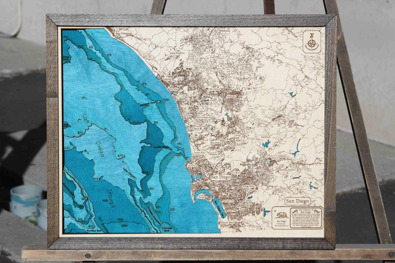 May include: A framed wooden map of San Diego with a blue and brown color scheme. The map shows the coastline and surrounding land. The map is framed in a dark wood frame. The map is titled 'San Diego' and includes a compass and a small text box with information about the city.