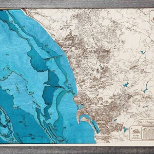 K&ouml;nnte beinhalten: Eine Holzkarte von San Diego, Kalifornien, mit einem blauen und braunen Farbschema. Die Karte zeigt die K&uuml;ste, die Stadt und die Umgebung. Die Karte ist in einem dunklen Holzrahmen gerahmt. Der Text "San Diego" ist in der unteren rechten Ecke der Karte gedruckt.