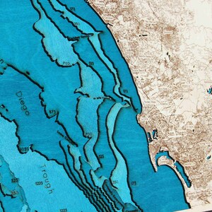 K&ouml;nnte beinhalten: Eine Holzkarte der K&uuml;ste von San Diego mit blauem Wasser und braunem Land. Die Karte zeigt Tiefenkonturen in Fu&szlig;, wobei der tiefste Punkt 1225 Fu&szlig; betr&auml;gt. Die Karte ist in einem dunklen Holzrahmen gerahmt.