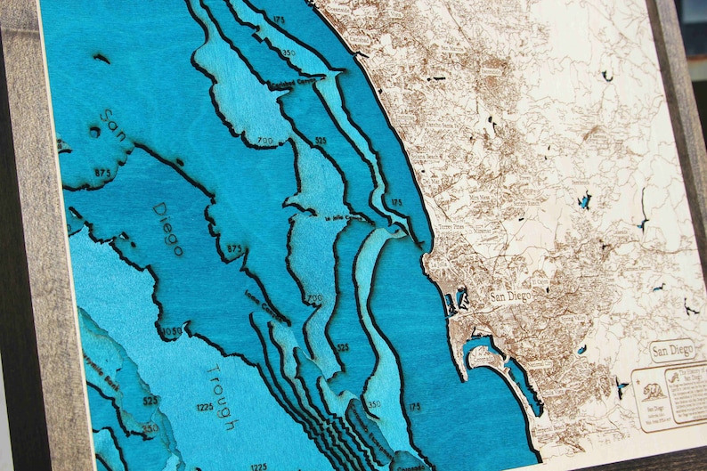 K&ouml;nnte beinhalten: Eine Holzkarte der K&uuml;ste von San Diego mit blauem Wasser und braunem Land. Die Karte zeigt Tiefenkonturen in Fu&szlig;, wobei der tiefste Punkt 1225 Fu&szlig; betr&auml;gt. Die Karte ist in einem dunklen Holzrahmen gerahmt.