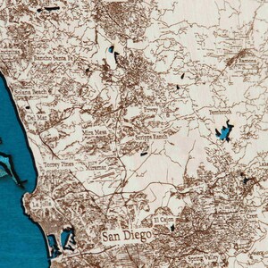 K&ouml;nnte beinhalten: Eine Holzkarte von San Diego, Kalifornien, mit den Stadtvierteln und der Umgebung in Braun umrandet. Die Karte ist auf einem blauen Hintergrund montiert, wobei das Meer und die umliegenden Wasserwege in Blau hervorgehoben sind.