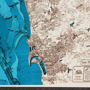 K&ouml;nnte beinhalten: Eine Holzkarte von San Diego, Kalifornien, mit blauem Meer und braunen Landmerkmalen. Die Karte ist in einem dunklen Holzrahmen gerahmt. Der Text "The History of San Diego" ist in der unteren rechten Ecke sichtbar.