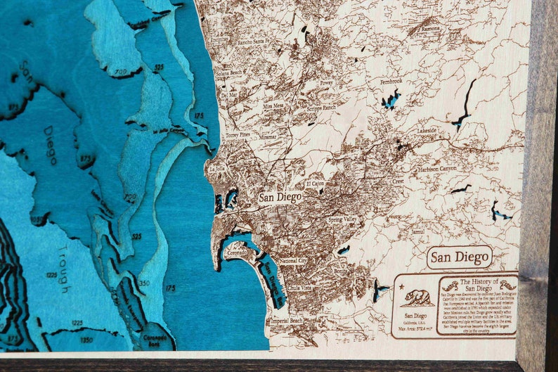 K&ouml;nnte beinhalten: Eine Holzkarte von San Diego, Kalifornien, mit blauem Meer und braunen Landmerkmalen. Die Karte ist in einem dunklen Holzrahmen gerahmt. Der Text "The History of San Diego" ist in der unteren rechten Ecke sichtbar.