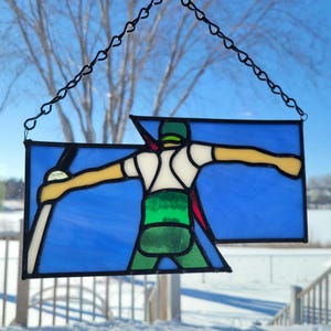 May include: A stained glass window hanging with a black chain. The window depicts a person in a green, white, and red outfit holding a ski pole. The background is blue.
