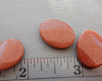 May include: Three oval-shaped, polished sandstone cabochons in shades of orange and brown. The stones are approximately 1 inch long and are arranged on a ruler.