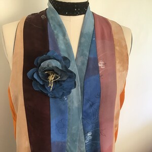 May include: A multi-colored silk scarf with a blue flower brooch. The scarf has stripes of orange, beige, brown, blue, and light blue. The flower brooch is made of fabric and has a gold wire center.