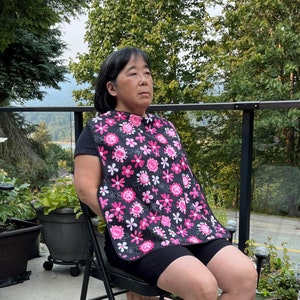 May include: A woman wearing a black and pink floral bib. The bib has a pink and white floral pattern with a black background. The woman is sitting on a black chair with a black railing behind her.