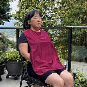 May include: A woman wearing a burgundy bib-style apron, sitting on a chair with a black metal frame. She is looking to the right of the camera.