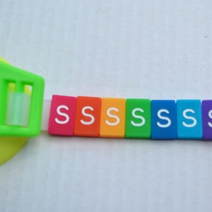 May include: A rainbow-colored set of letter tiles spelling out the word "SSSSSSS" in white letters. The tiles are attached to a white strap.