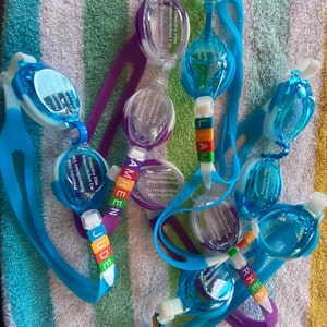May include: A close-up of a group of colorful swimming goggles with names written on the straps. The goggles are on a striped towel. The names on the straps are AMREEN, JUDE, LIAM, RHEA.