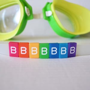Puede incluir: Un conjunto de fichas de letras de colores arcoíris que deletrean la palabra "BBBBBBB" en letras blancas. Las fichas están sobre una superficie blanca delante de un par de gafas de natación verdes y amarillas.