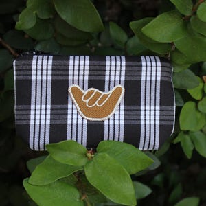 May include: A black and white plaid zippered pouch with a brown hand gesture patch. The patch is a stylized hand with the fingers curled in a gesture of greeting or good luck.